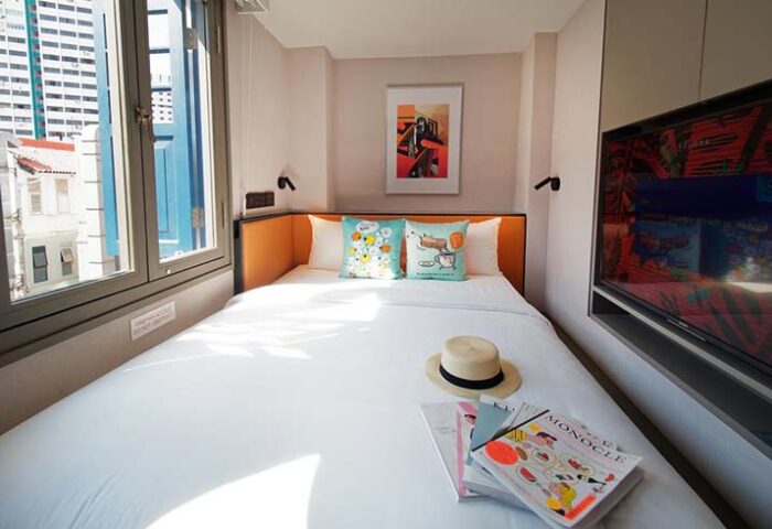 Hotel Soloha @ Chinatown gay-friendly hotel room. Bed with pillows, hat, magazines, and a TV. Window view of Singapore buildings.