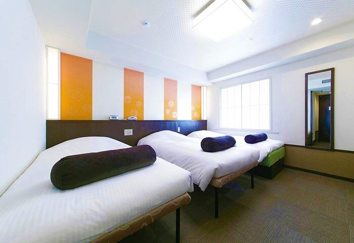 Hotel Sunlite Shinjuku's gay-friendly room with three beds, brown headboard, and orange wall art. A mirror reflects the hallway.