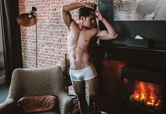 Gay-friendly Hotel V Frederiksplein room. Shirtless man in underwear poses by fireplace. Brick wall, chair, and decor in Amsterdam hotel.
