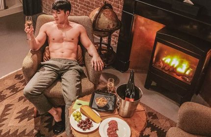 Hotel V Frederiksplein gay-friendly hotel room: Shirtless man relaxing with champagne, cheese, and salami. Fireplace and globe visible.