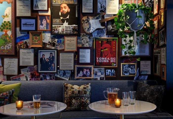 Hotel Ziggy Los Angeles's gay-friendly vibe. Wall decor with music memorabilia, Nipsey Hussle poster, and Tone-Loc flyer. Tables with drinks.