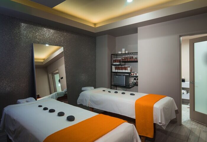 Hotel Zoso gay-friendly hotel spa. Two massage tables with orange accents, black stones, and a reflective wall. Shelves with spa products.