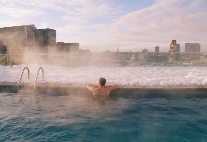 Gay-friendly Hyatt Centric Montreal: Man relaxing in rooftop pool, snow-covered edge, city skyline view. Ideal for gay travel, Montreal.