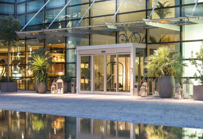 INNSiDE Milano Torre Galfa entrance. Gay-friendly hotel with modern design, glass facade, plants, and decorative lanterns. Upscale Milan lodging.