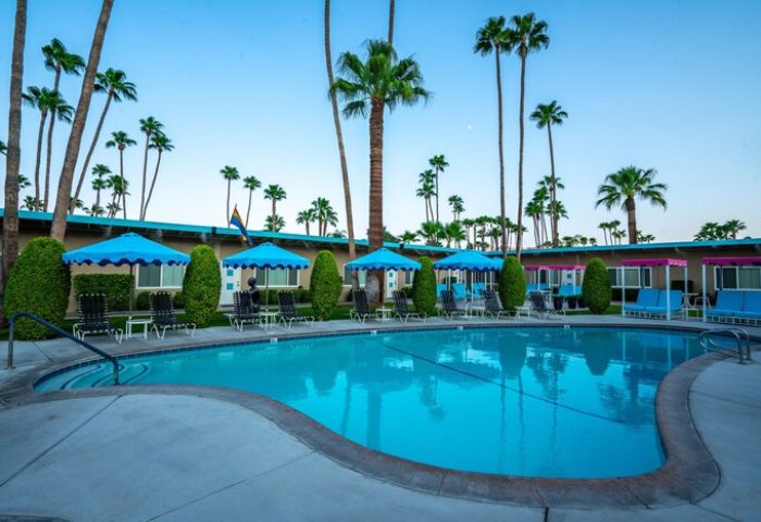 Pool view at INNdulge, an iconic gay men's resort in Palm Springs. Blue umbrellas, lounge chairs, and palm trees create a relaxing, gay-friendly vibe.