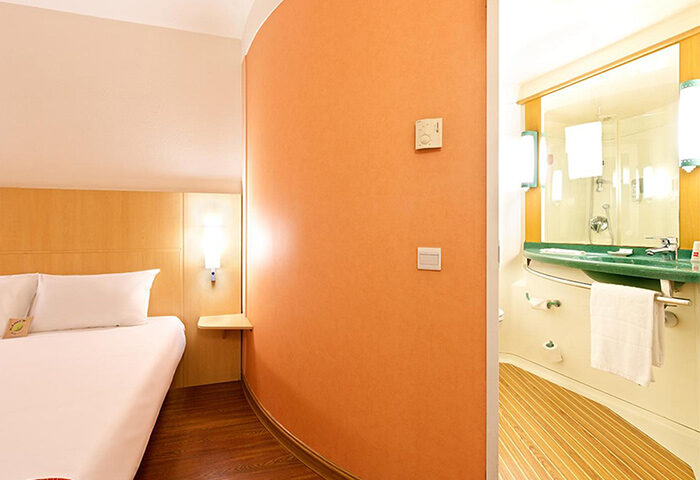 Ibis Beijing Dongdaqiao Hotel room. Gay-friendly hotel featuring a bed with white linens, orange wall, and view into the bathroom.