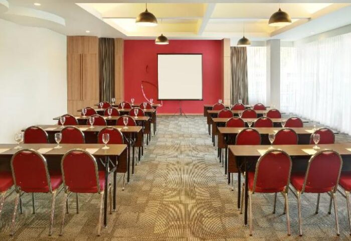 Ibis Budget Bali Seminyak meeting room setup. Rows of tables and red chairs face a projector screen. Gay-friendly hotel for events.