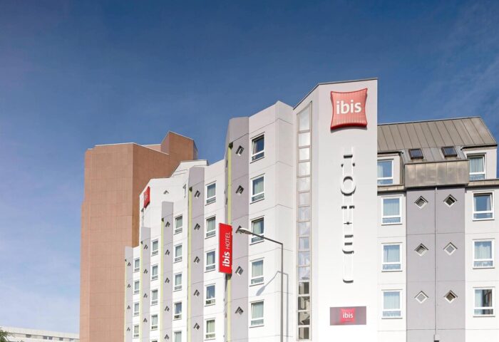 Ibis Hotel Koeln Centrum exterior. Gay-friendly hotel in Cologne, Germany. Modern building with Ibis logo and red banners.