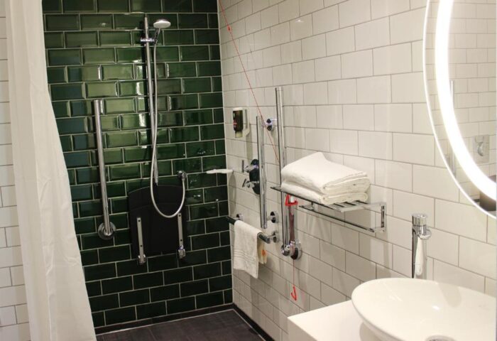 Ibis Styles London Southwark Hotel bathroom. Accessible shower with seat, grab bars, and white and green tiling. Gay-friendly hotel.