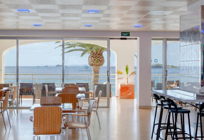 Gay-friendly hotel in Ibiza Playa: Interior view of dining area with ocean view. Tables, chairs, bar stools, and a palm tree visible.