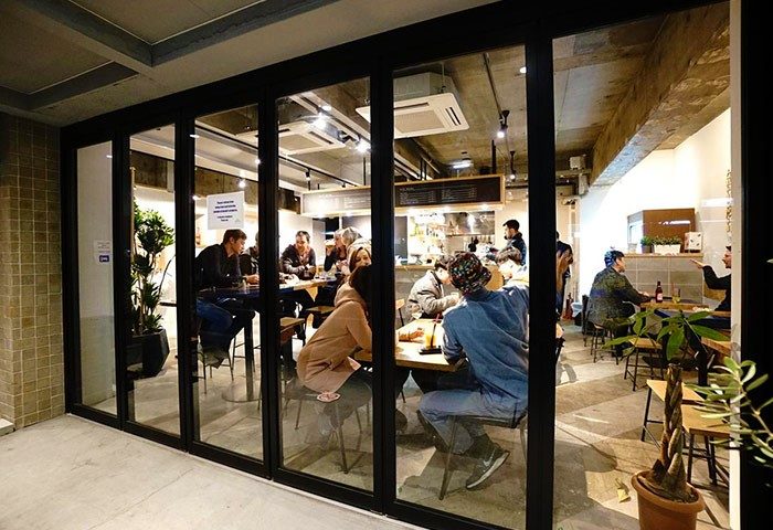 Imano Tokyo Hostel's gay-friendly hotel cafe. People are seated at tables inside. The cafe is visible through a glass wall.