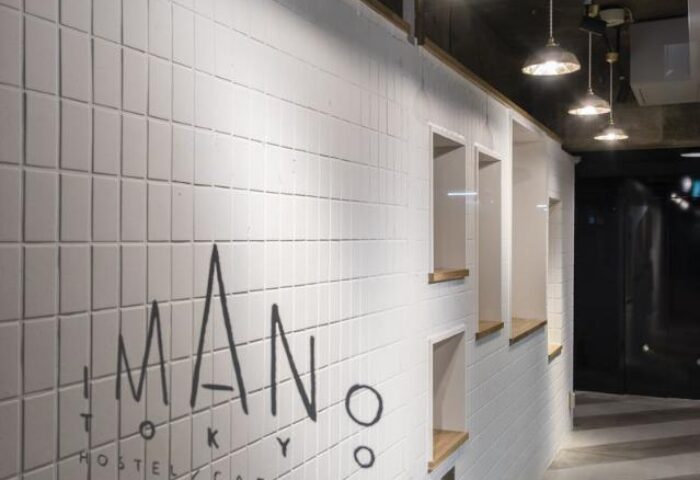 Imano Tokyo Hostel interior. White tiled wall with Imano Tokyo Hostel Cafe & Bar logo. Gay-friendly hotel with modern design elements.