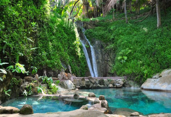 Imanta Resorts Punta De Mita gay-friendly hotel view. Waterfall flowing into a clear pool, lush greenery. Perfect for gay men's travel.