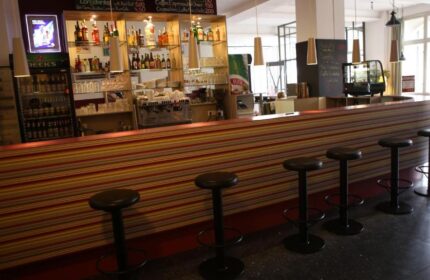 Bar area at Industriepalast Hostel, a gay-friendly hotel in Berlin. Stools line the striped bar. Shelves display bottles and glasses.