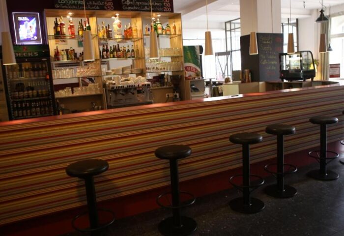 Bar area at Industriepalast Hostel, a gay-friendly hotel in Berlin. Stools line the striped bar. Shelves display bottles and glasses.