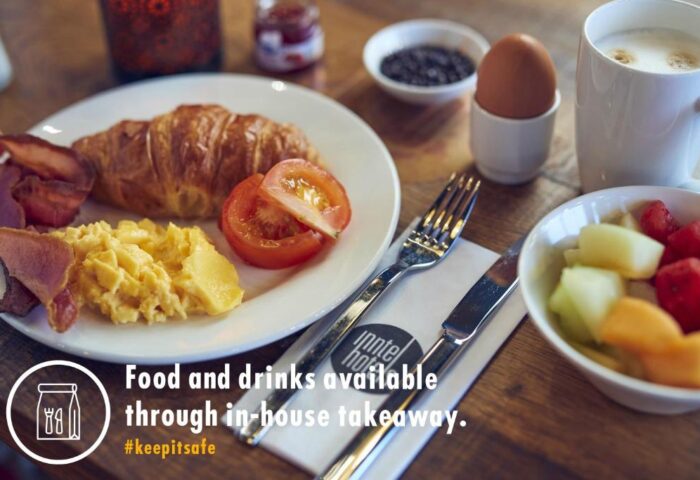 Breakfast at Inntel Hotels Amsterdam Centre, a gay-friendly, pet-friendly hotel. Croissant, eggs, bacon, fruit, and coffee on a wooden table.