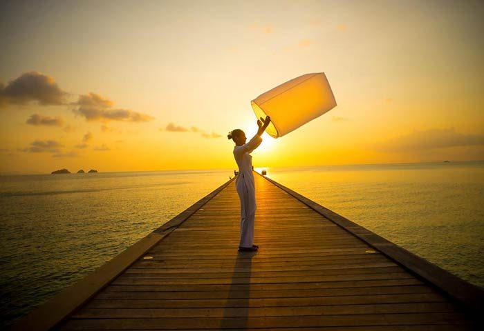 Sunset at InterContinental Koh Samui Resort, a gay-friendly hotel. Woman on pier releases lantern into the golden sky. Romantic Thailand getaway.