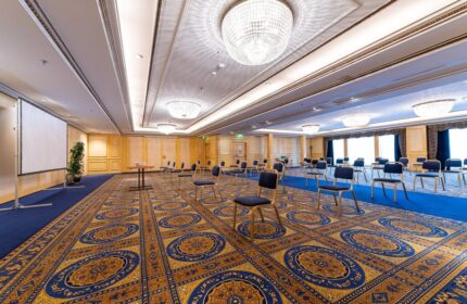 InterContinental Wien's ballroom, a gay-friendly hotel, features a projector screen, chandeliers, and chairs spaced apart on a patterned carpet.