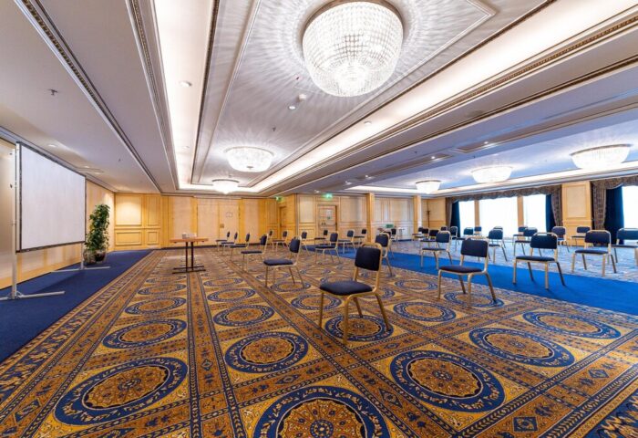 InterContinental Wien's ballroom, a gay-friendly hotel, features a projector screen, chandeliers, and chairs spaced apart on a patterned carpet.