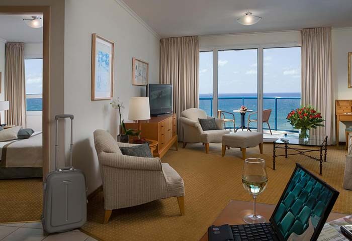 Isrotel Tower Gay-Friendly Hotel room. Tel Aviv luxury suite with ocean view, balcony, seating area, laptop, and suitcase. Perfect for gay travel.