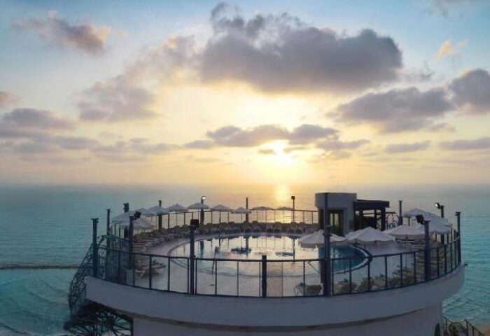 Rooftop pool at Isrotel Tower, a gay-friendly hotel. Chairs and umbrellas surround the pool with a scenic ocean view at sunset.
