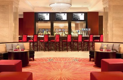 Bar at the gay-friendly JW Marriott Chicago hotel. Red chairs line the bar, with TVs above. Seating areas flank a patterned carpet.