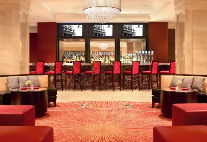 Bar at the gay-friendly JW Marriott Chicago hotel. Red chairs line the bar, with TVs above. Seating areas flank a patterned carpet.