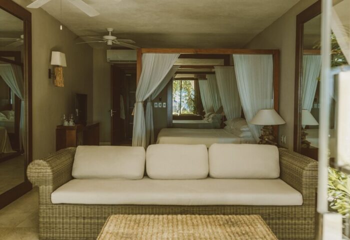 Jashita Hotel room: Wicker couch and table in front of a four-poster bed with white curtains. Gay-friendly hotel room with neutral tones.