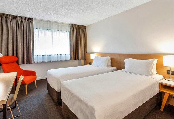 Jasper Boutique Hotel gay-friendly room. Two single beds with white linens, bedside table, lamp, orange chair, and window with curtains.