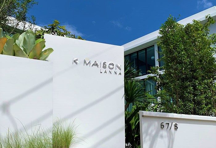 K Maison Lanna Boutique Hotel entrance. Gay-friendly hotel architecture with white walls, greenery, and clear blue sky. Modern design.