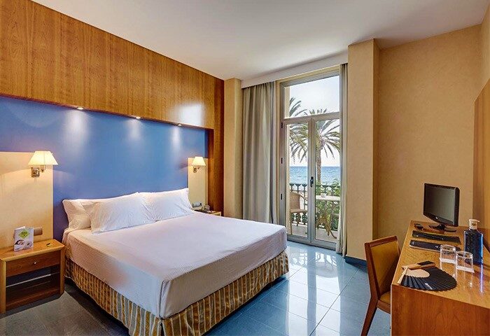 Kalma Sitges Hotel bedroom with balcony, sea view. Gay-friendly hotel in Sitges, Spain. Room features bed, desk, & ocean view. Palm tree outside.
