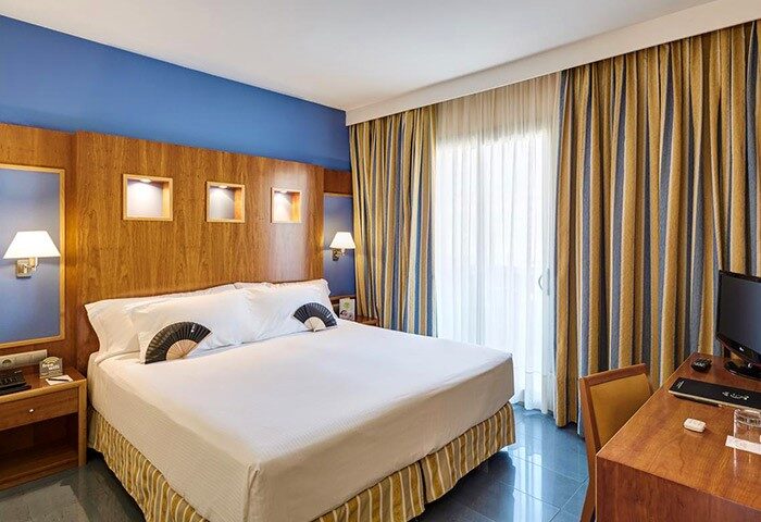 Kalma Sitges Hotel gay-friendly room. Double bed with fans, blue wall, wood headboard, desk, and striped curtains. Sitges gay travel.
