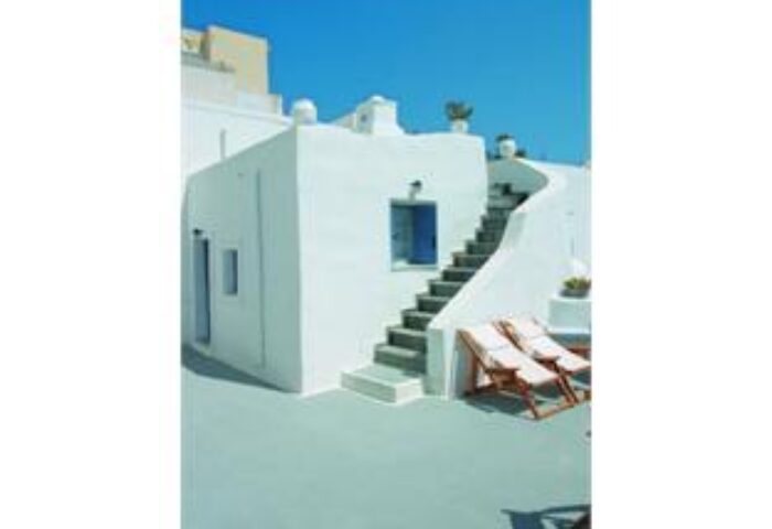 Gay-friendly Kamares Apartments hotel view. White building with blue door, stairs, and lounge chairs. Ideal for gay men travel in Greece.