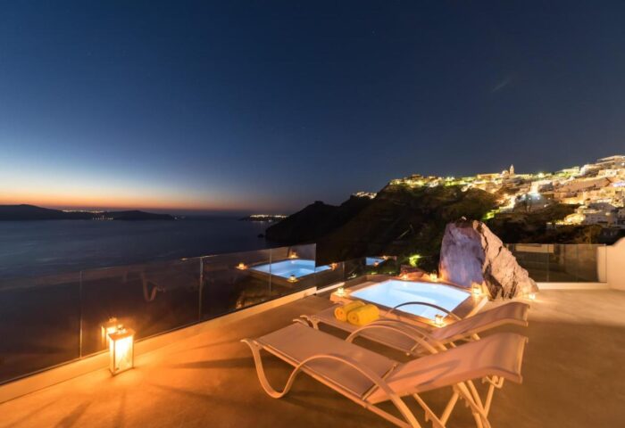 Keti Hotel, a gay-friendly hotel. Jacuzzi and lounge chairs on a terrace at night overlooking the sea and city lights in Santorini.