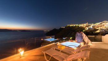 Keti Hotel, a gay-friendly hotel. Jacuzzi and lounge chairs on a terrace at night overlooking the sea and city lights in Santorini.