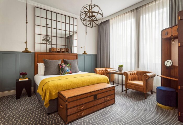 Kimpton Clocktower Hotel room: bed with yellow throw, leather trunk, chairs, & grid mirror. Gay-friendly hotel design with stylish details.
