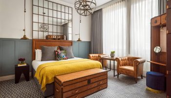 Kimpton Clocktower Hotel room: bed with yellow throw, leather trunk, chairs, & grid mirror. Gay-friendly hotel design with stylish details.