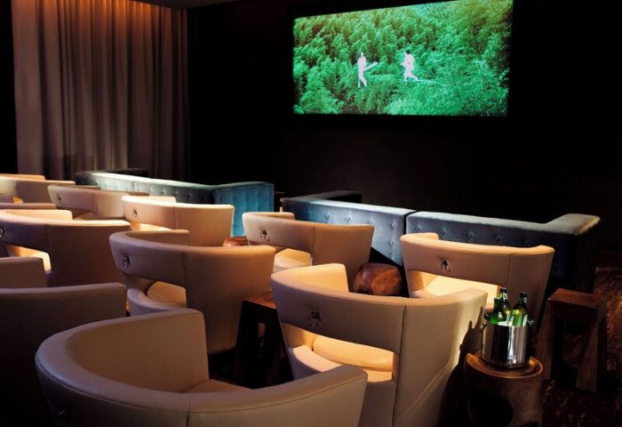 Kimpton Hotel Eventi gay-friendly cinema. Theater seating with curved chairs, couches, and a large screen showing men running in a forest.