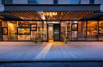 Kimpton Hotel Eventi, gay-friendly hotel entrance. Glass facade, lights, and plants. Welcoming ambiance for gay travelers. NYC hotel.