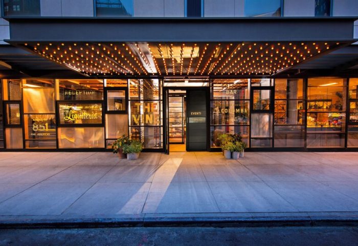 Kimpton Hotel Eventi, gay-friendly hotel entrance. Glass facade, lights, and plants. Welcoming ambiance for gay travelers. NYC hotel.