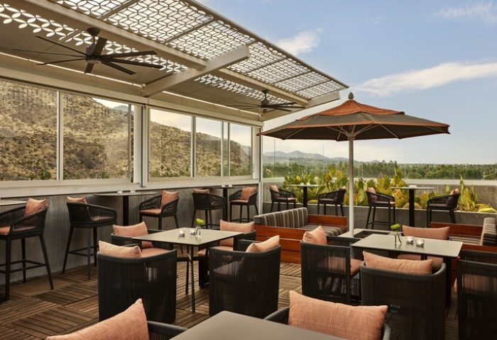 Outdoor dining at gay-friendly Kimpton Rowan Palm Springs Hotel. Rooftop bar with mountain views, tables, chairs, and umbrellas. Stylish hotel.