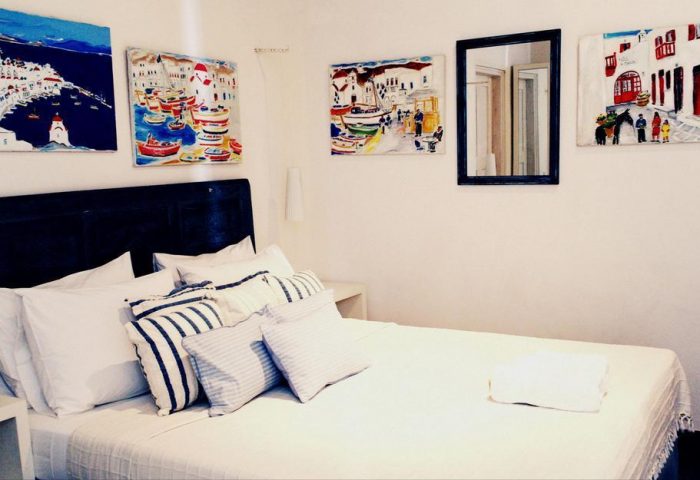 La Maison Kalogera gay-friendly hotel room. Bed with white linens, paintings of Mykonos, Greece, and a mirror on the wall.