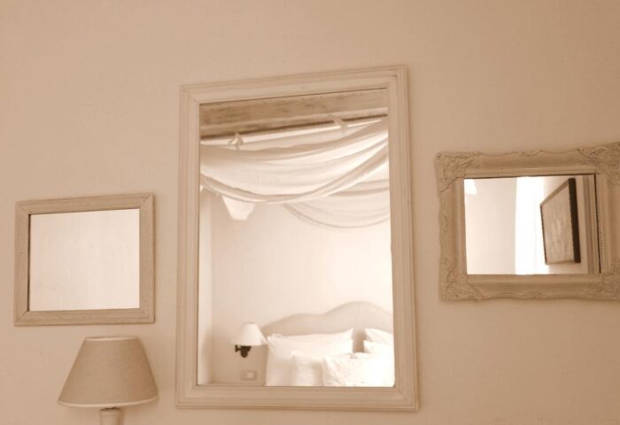 La Maison Kalogera, gay-friendly hotel detail. Mirrors reflect a bedroom with white linens and draped fabric. Lamp on bedside table. Calm, serene.
