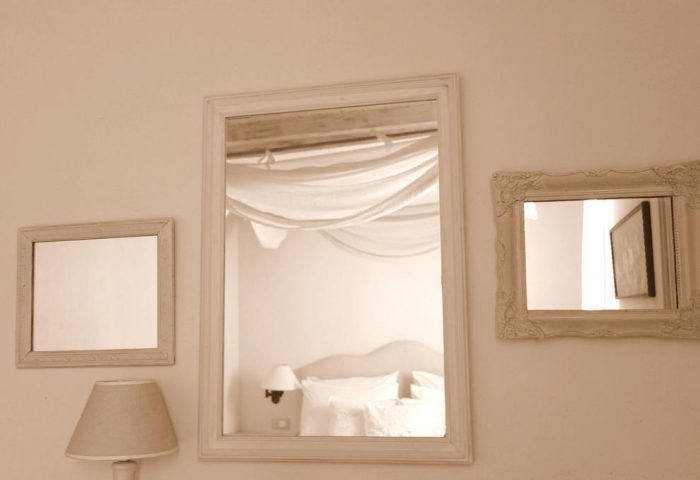 La Maison Kalogera, gay-friendly hotel detail. Mirrors reflect a bedroom with white linens and draped fabric. Lamp on bedside table. Calm, serene.