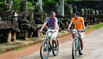 Gay men biking near La Residence d'Angkor, a Belmond Hotel, Siem Reap. Stone statues line the road. Gay-Friendly Hotel in Cambodia.
