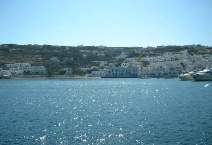Mykonos guesthouse view from the sea. Traditional white buildings on a hill, yachts in the water. Gay-friendly hotel, La Veranda of Mykonos.
