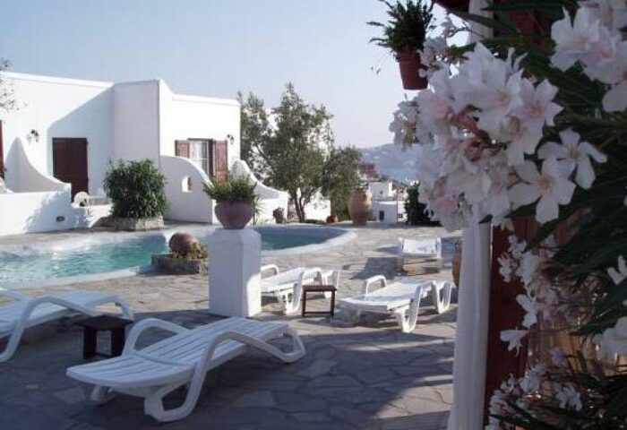 Poolside view at La Veranda of Mykonos Traditional Guesthouse, a gay-friendly hotel. White buildings, sunbeds, and flowers.