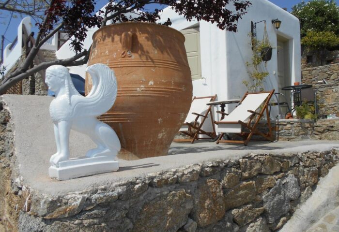 La Veranda of Mykonos Traditional Guesthouse view. Sphinx statue, large pot, deck chairs, and whitewashed building. Gay-friendly hotel detail.