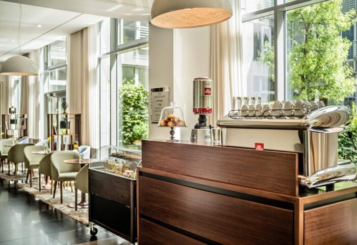Le Méridien Munich gay-friendly hotel cafe. Illy coffee machine, pastries, and seating area. Modern interior design with natural light.