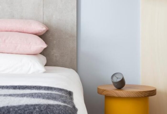 Leman Locke Hotel detail: Bed with pink pillows, grey blanket, yellow nightstand, and black lamp. Gay-friendly hotel room interior.